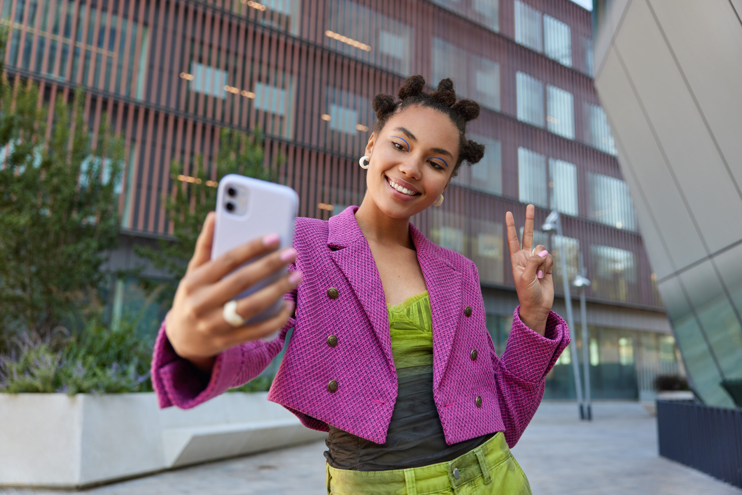 Attractive girl in fashionable outfit creats influence content shows peace sign at smartphone front camera poses for selfie smiles gladfully poses at urban place spends vacation at big city.